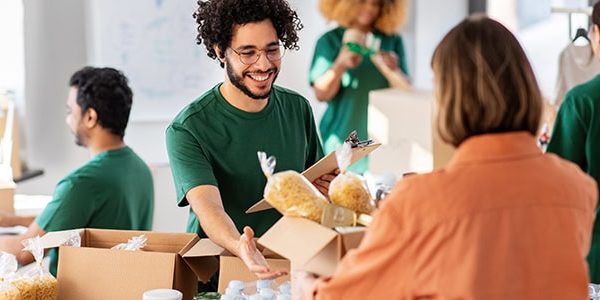 charity, donation and volunteering concept - happy smiling male volunteer with clipboard and woman taking box of food at distribution or refugee assistance center